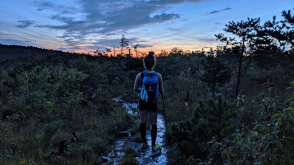 girl with bun and blue backpack walking on a wet mountain trail at sunrise, the water on the trail is reflecting the sunrise