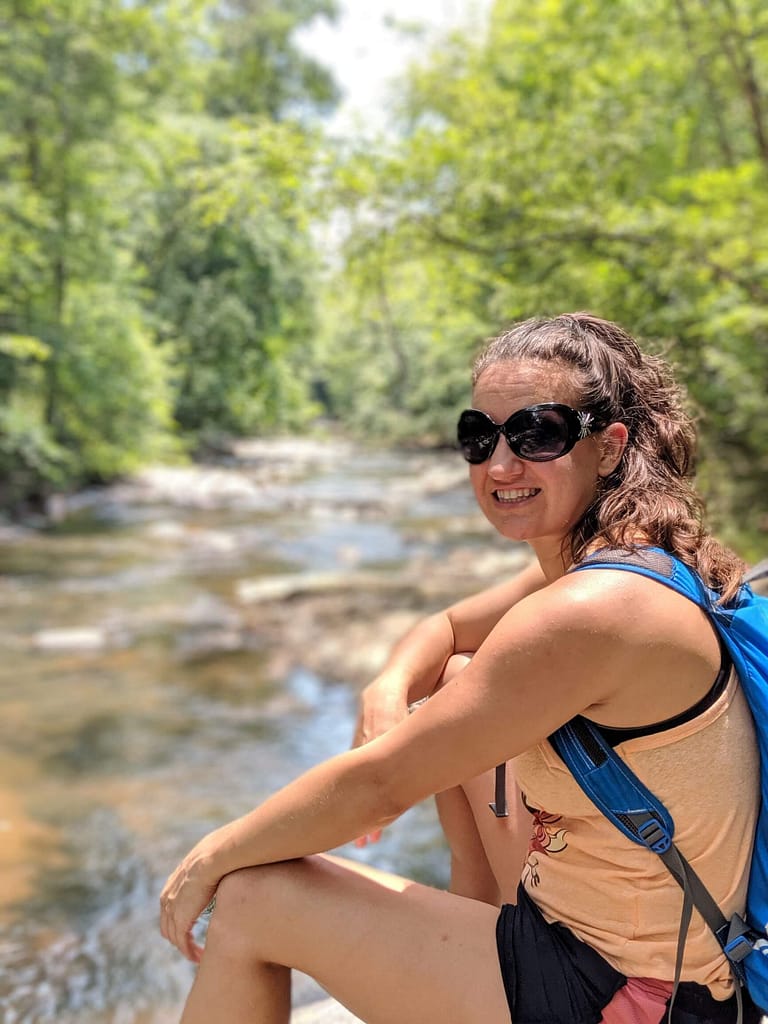 Girl with sunglasses and orange shirt wearing a backpack and sitting in front of a creek smiling